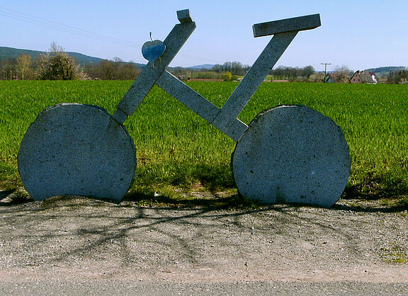 Sehr großes Fahrrad aus Stein am Wegesrand. Im Hintergrund große grüne Weide.