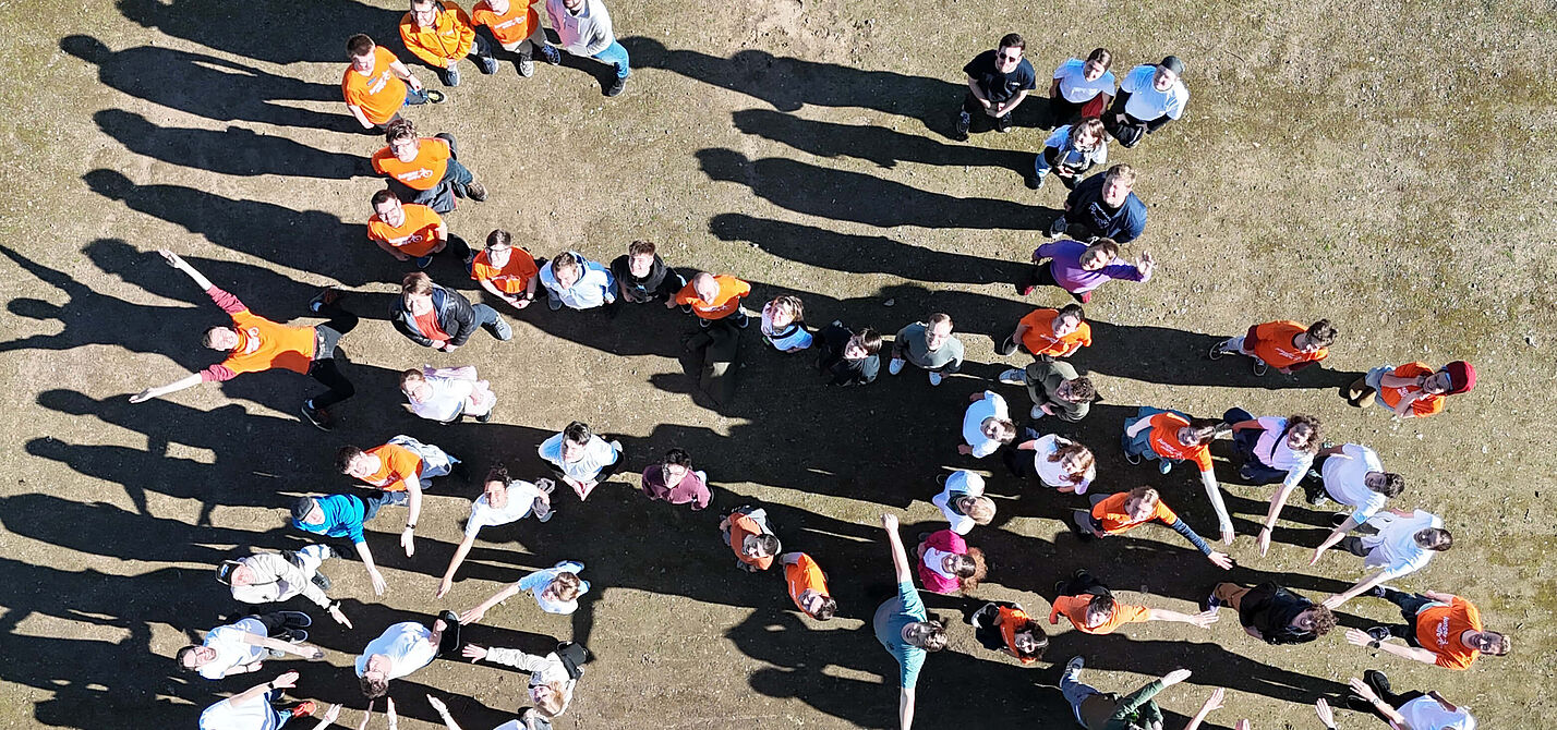 Gruppe von Menschen steht auf einem Platz und bildet gemeinsam die Form eines Fahrrads. Die Personen tragen teils orangefarbene, teils weiße oder bunte Oberteile. Das Bild wurde aus der Vogelperspektive aufgenommen.