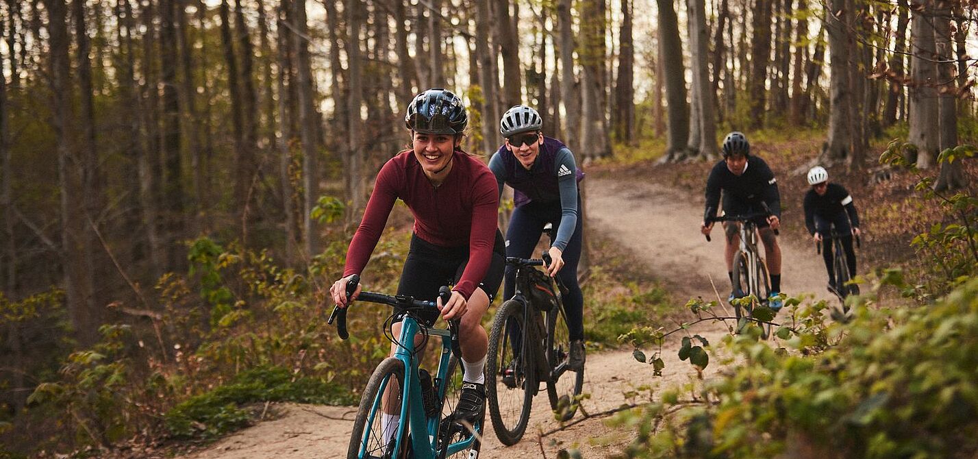 Mit Gravelbikes abseits des Asphalts unterwegs. Eine Gruppe von Frauen und Männern fahren mit Gravelbikes hintereinander auf einem Waldweg.