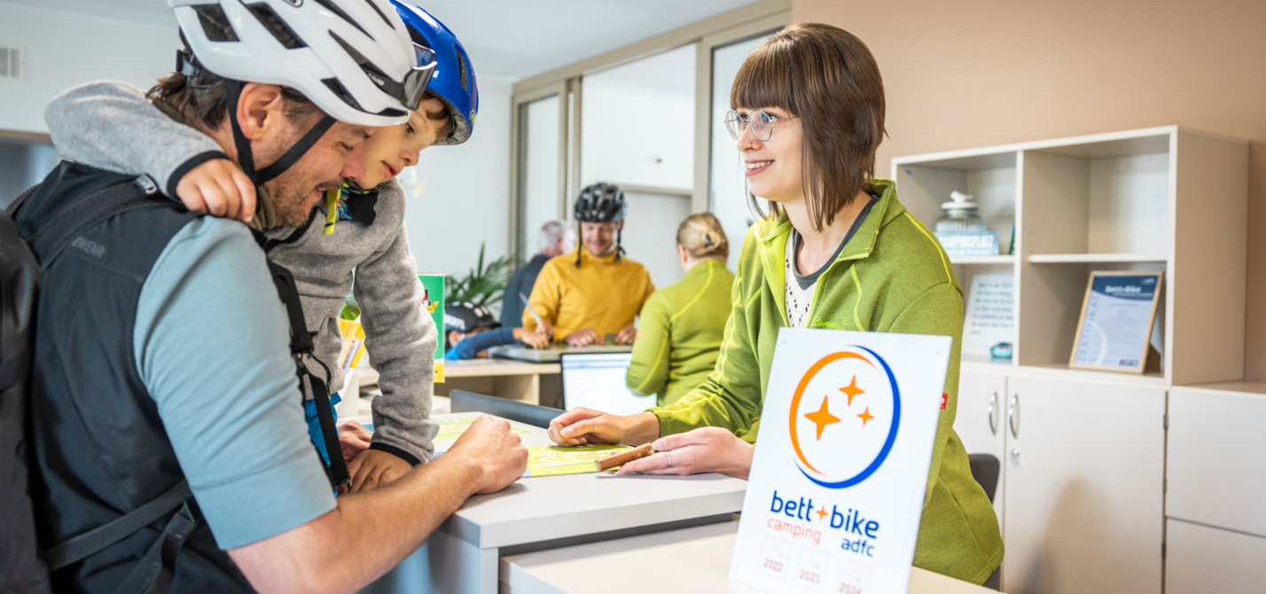 Bett+Bike - Rezeption Camping Ein Vater mit Kind auf dem Arm informiert sich an einer Rezeption eines Campingplatzes, beide tragen Fahrradhelme. Im Vordergrund steht ein Bett+Bike-Camping-Schild.