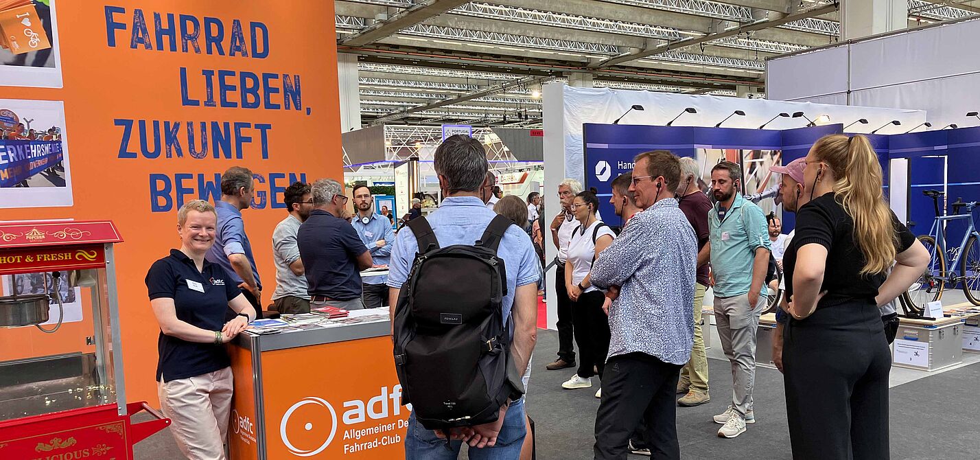 Ein Messestand des ADFC in einer großen Halle. Vor einer orangen Rückwand mit dem blauen Slogan „Fahrrad lieben, Zukunft bewegen“ steht eine lächelnde Frau im dunkelblauen ADFC-Shirt an einer Theke. Mehrere Menschen stehen davor und dahinter in Gruppen zusammen und unterhalten sich. Im Vordergrund links ist eine rote Popcornmaschine zu sehen.